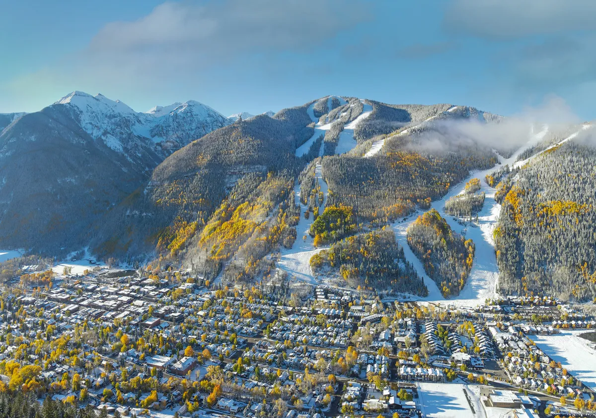 A sweeping view of snow-covered mountains surrounding Telluride, with leafy trees, ski slopes, and the welcoming town nestled below. The scene hints at both adventure on the slopes and cozy returns to town life.