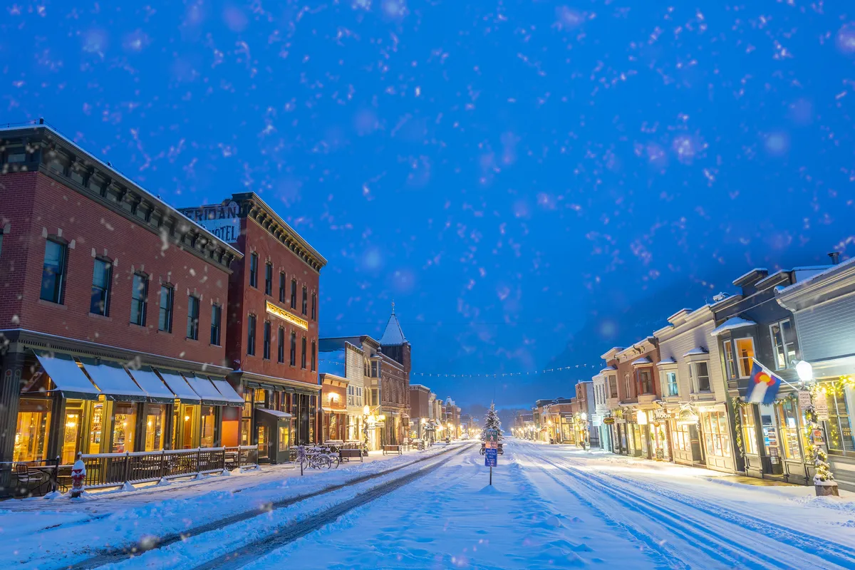 A snow-dusted street lined with historic buildings, festive holiday lights, and a large Christmas tree. People are out shopping and enjoying the magic of winter in the heart of Telluride.