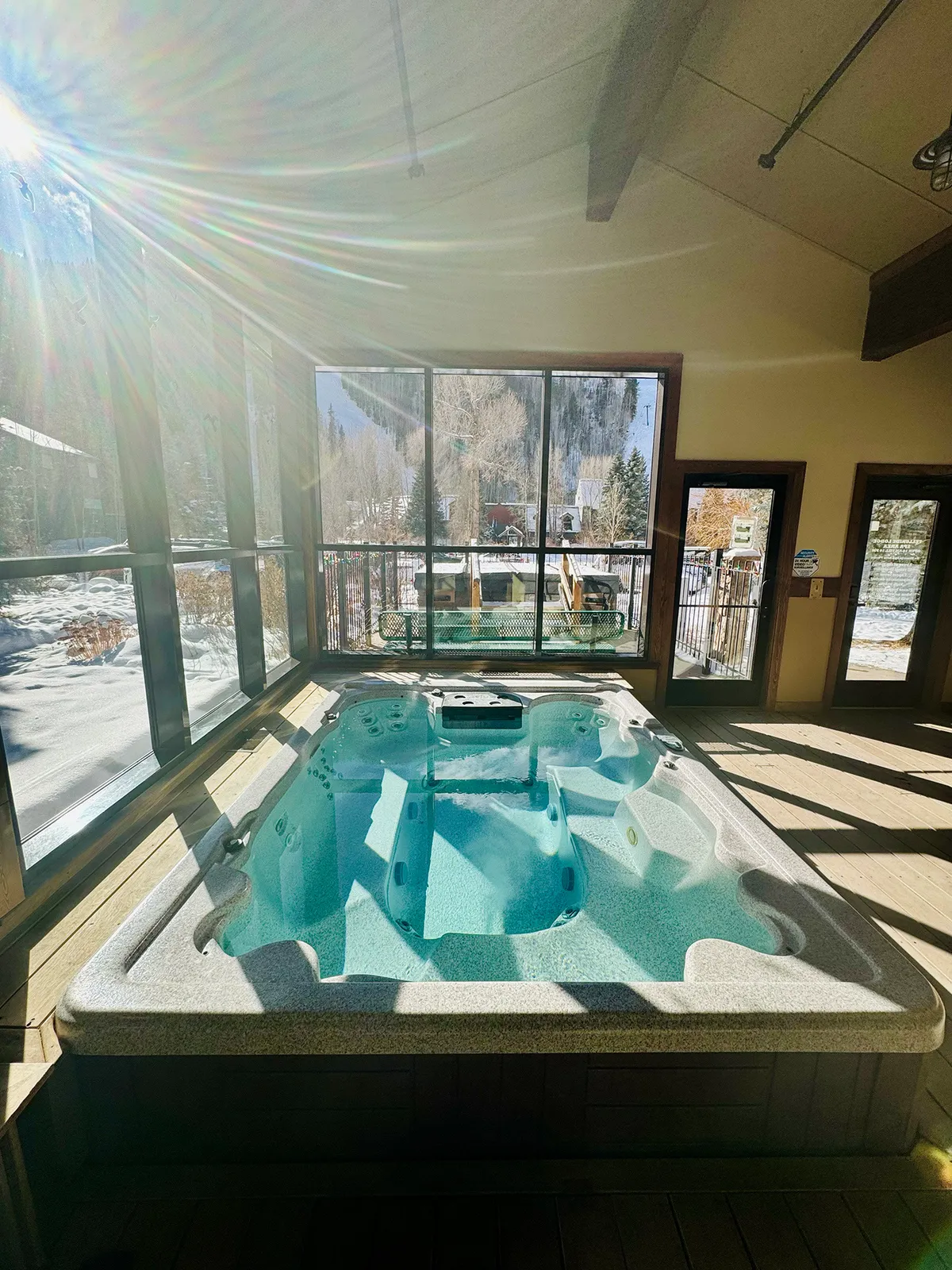 This image captures a communal hot tub set within a bright interior, featuring large windows that reveal mountain views and natural light pouring in. Wooden beams and an inviting, social atmosphere make the space ideal for friends or family to unwind together after a day outdoors.