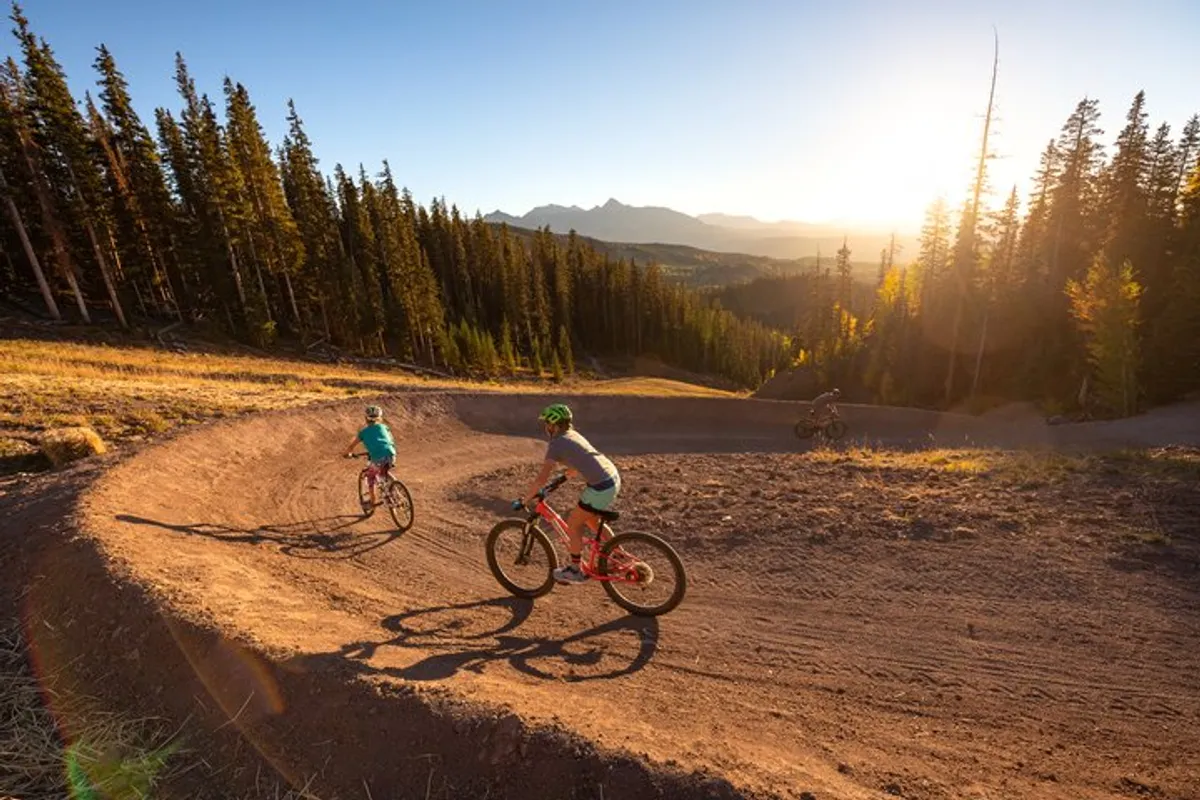Two mountain bikers speed along a curved dirt trail flanked by tall pine trees, with a dramatic mountain range and a sunset sky in the distance. The scene is energetic, highlighting both the thrill of the ride and the natural beauty of Telluride’s varied terrain.