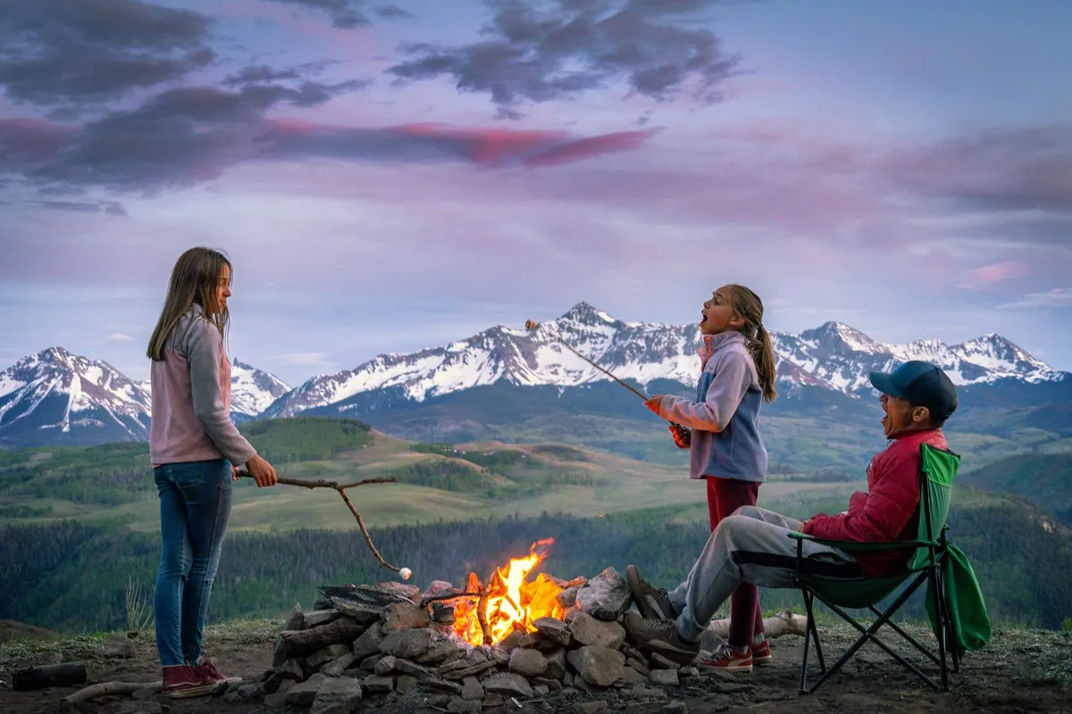 A group gathers around a glowing campfire with children roasting marshmallows, set against a scenic mountain backdrop. Lush grass and open space invite play, while the fire pit and seating offer a hub for evening stories and togetherness.