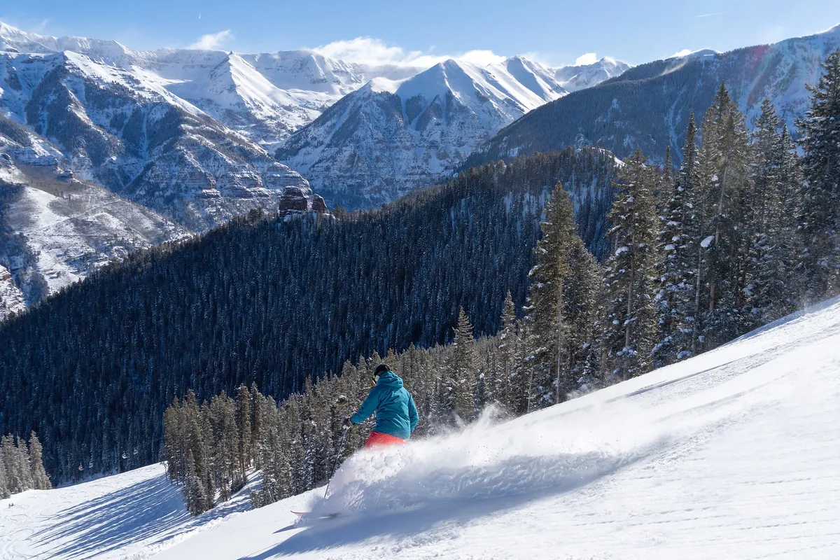 A skier carves down a pristine, snow-covered slope with stunning, rugged mountains towering in the background. The fresh powder and clear blue sky evoke the exhilaration of winter sports, and the image captures the energy and scale of Telluride's world-class ski terrain.