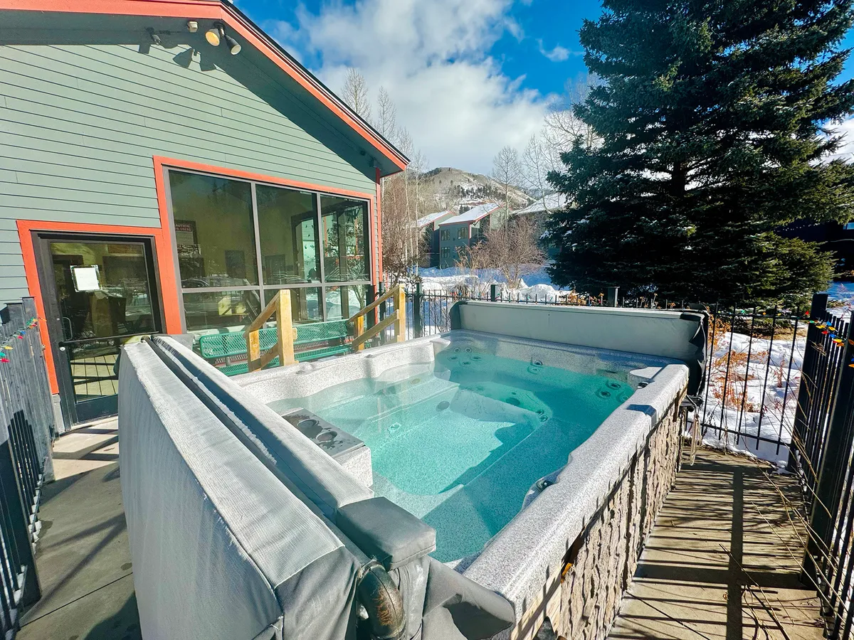 An exterior view showing a hot tub adjacent to a home, surrounded by trees and set against a backdrop of mountains. The scene evokes a sense of relaxation, connection to nature, and the possibility of hosting outdoor gatherings like barbecues.