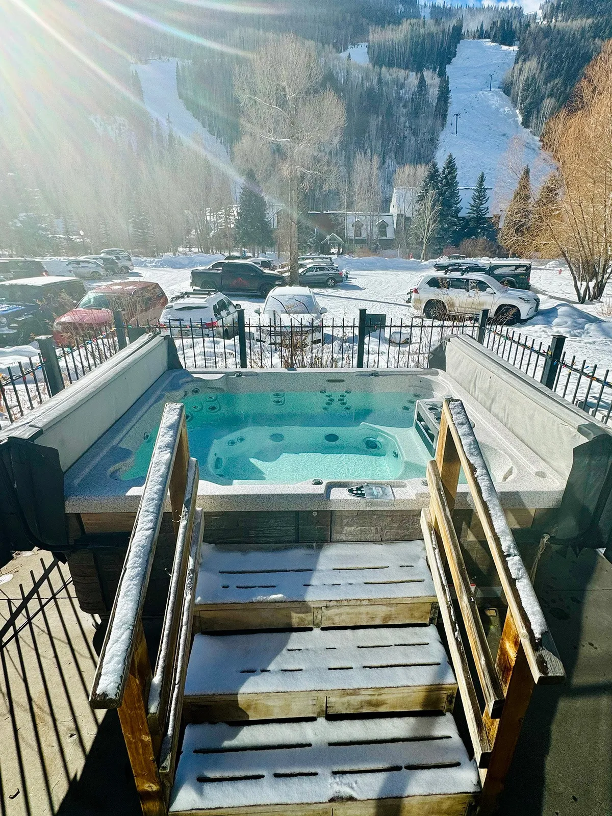 An outdoor hot tub sits nestled among snow, with steam rising into the crisp air. Mountain views sprawl in the background, and the space feels inviting for both relaxation and conversation. Steps and surrounding deck create a communal space ideal for gathering after a day outdoors.