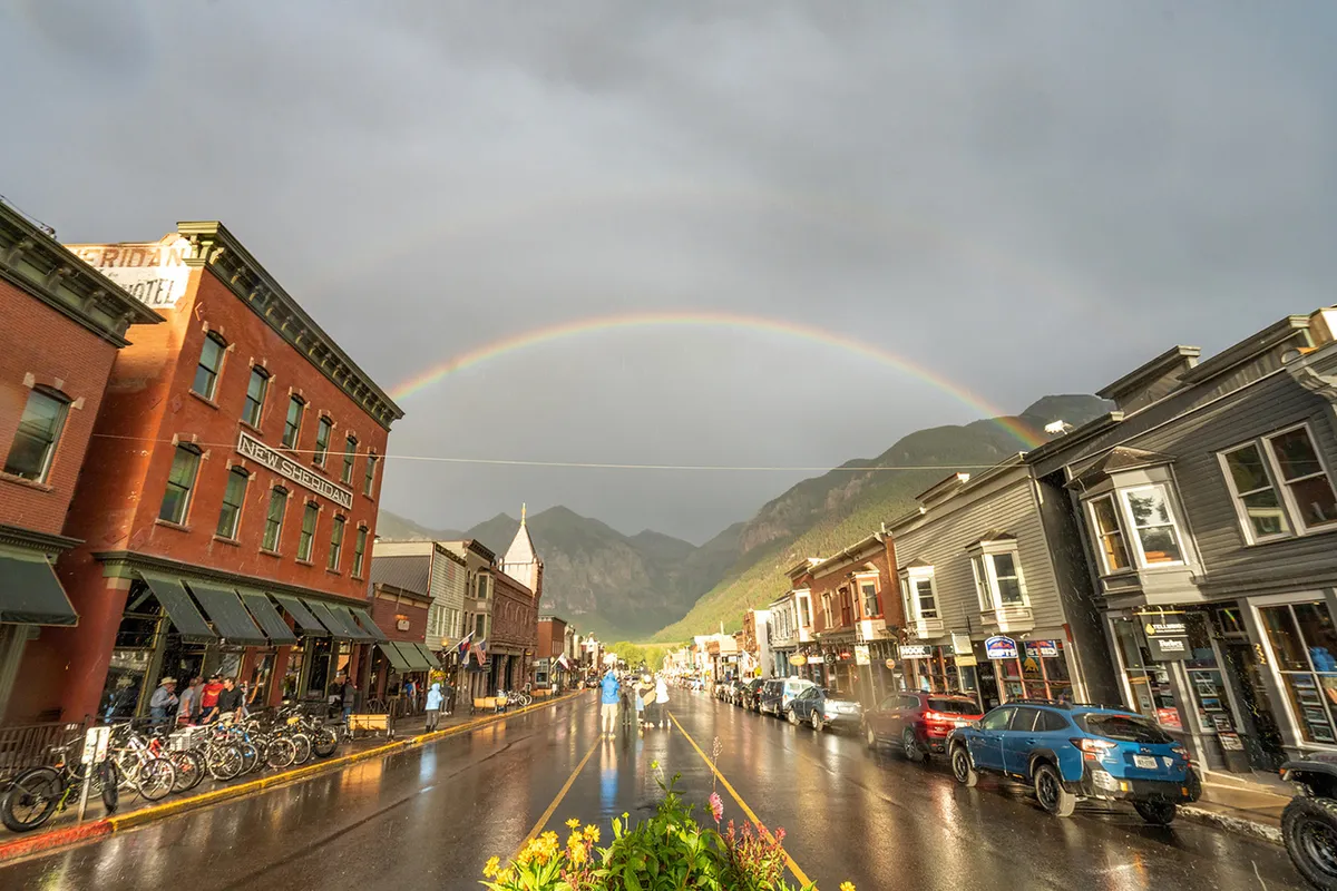 A vibrant street in Telluride featuring historic buildings, a rainbow arching over wet pavement, and distant mountain views. The scene is bustling yet charming, with signs of shopping, dining, and exploration all within easy reach.