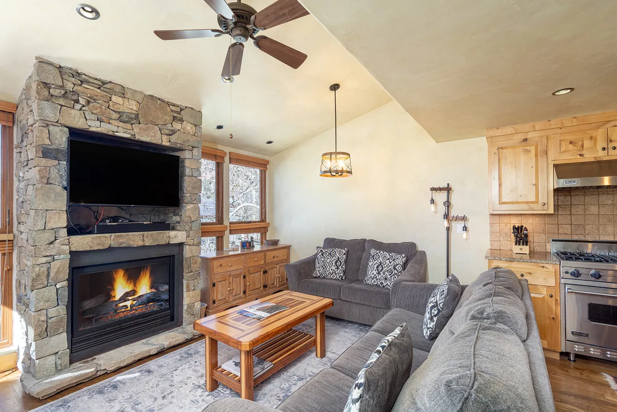 The image features a sunlit great room with a beautiful stone fireplace as the centerpiece, a plush comfortable couch, a rustic wooden coffee table, and expansive windows that flood the space with natural light. The inviting setting is perfect for relaxing or gathering with friends and family after a day of mountain adventures.