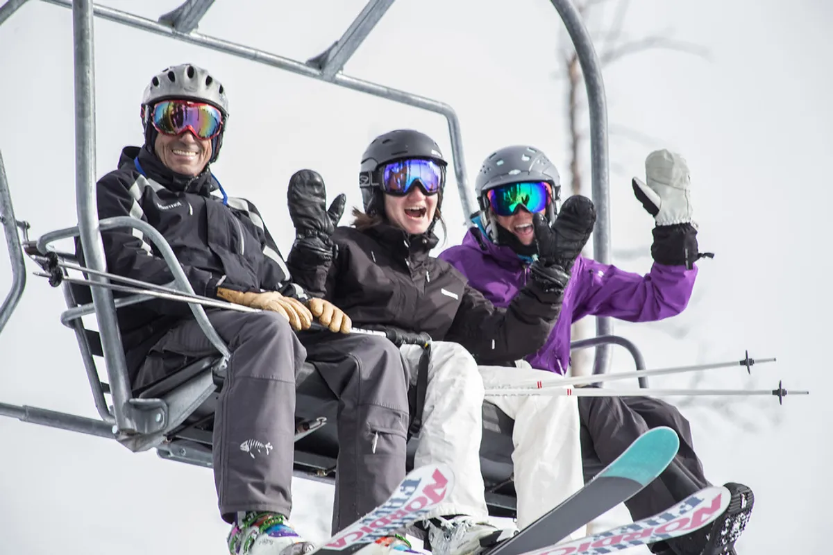 Three skiers, bundled in winter gear, ride a chairlift above a snowy landscape. The snowy slopes and cheerful mood evoke the thrill and camaraderie of Telluride's all-season outdoor adventures.