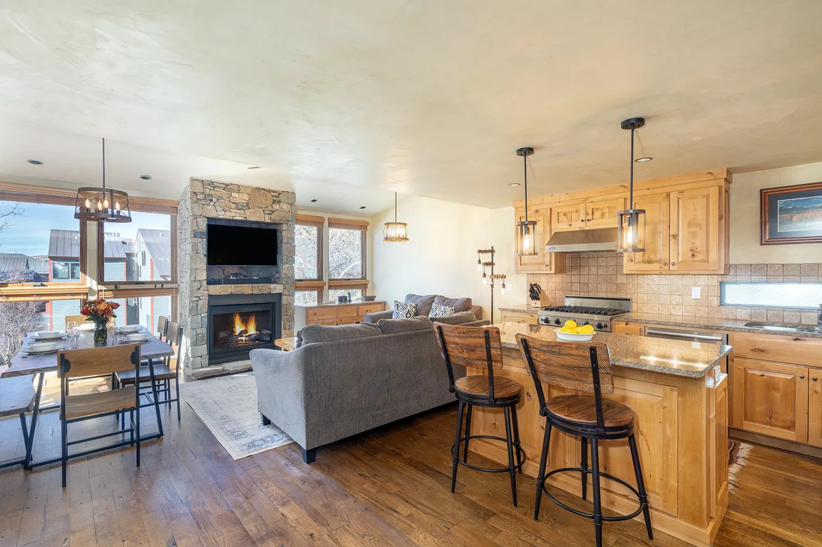 A warm, open-concept space featuring a stylish fireplace, a spacious kitchen island, a dining table, and expansive windows. Sunlight streams through the large windows, creating a welcoming, airy atmosphere that connects the living, dining, and kitchen areas.