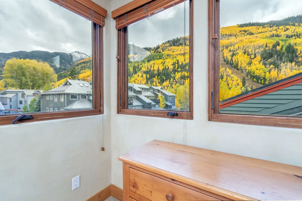 A cozy interior living area features large windows framing stunning mountain views, with sunlight spilling across wooden furniture. The scene invites guests to relax, read, or gather indoors while still feeling connected to nature outside.
